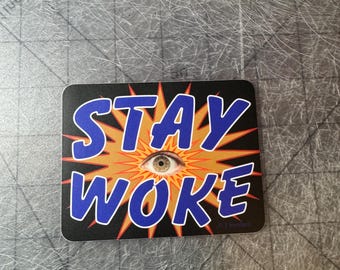 Puede incluir: Una pegatina negra y naranja con el texto "STAY WOKE" en letras azules. Un ojo grande con iris marrón está en el centro de la pegatina.
