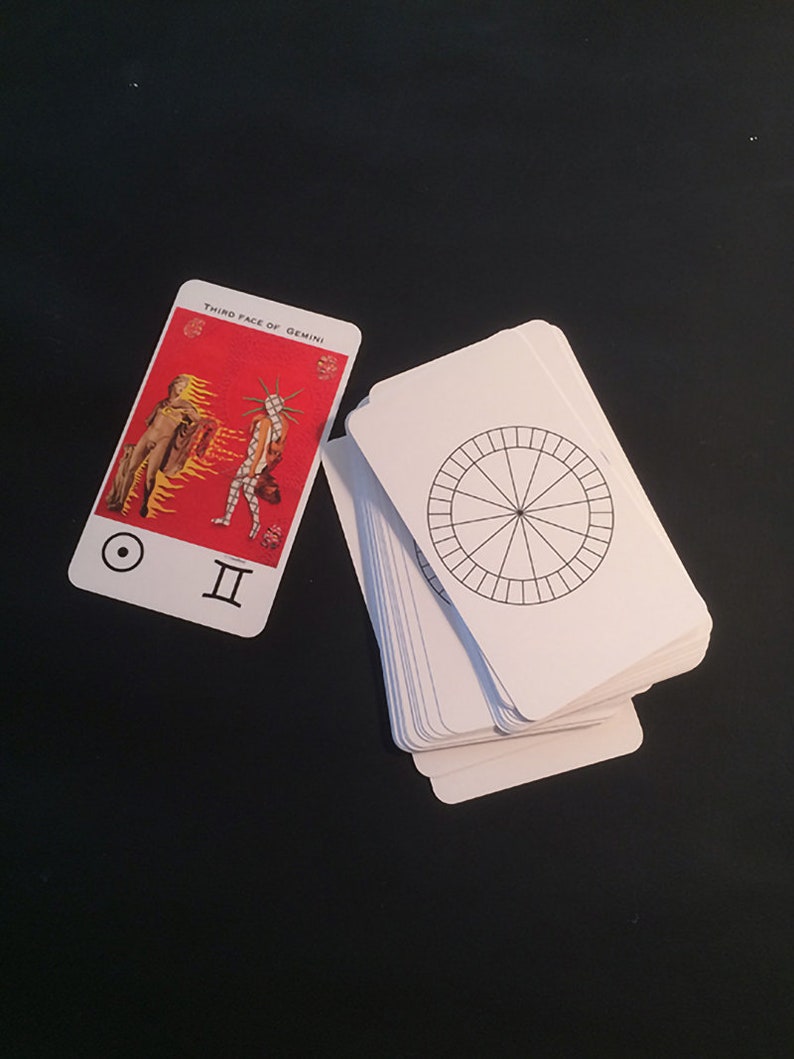May include: A tarot card with a red background and two figures, one in gold and one in a colorful costume. The card is labeled "Third Face of Gemini" and has the symbol for the sun and the Roman numeral II. A stack of blank tarot cards with a circle divided into sections is in the background.
