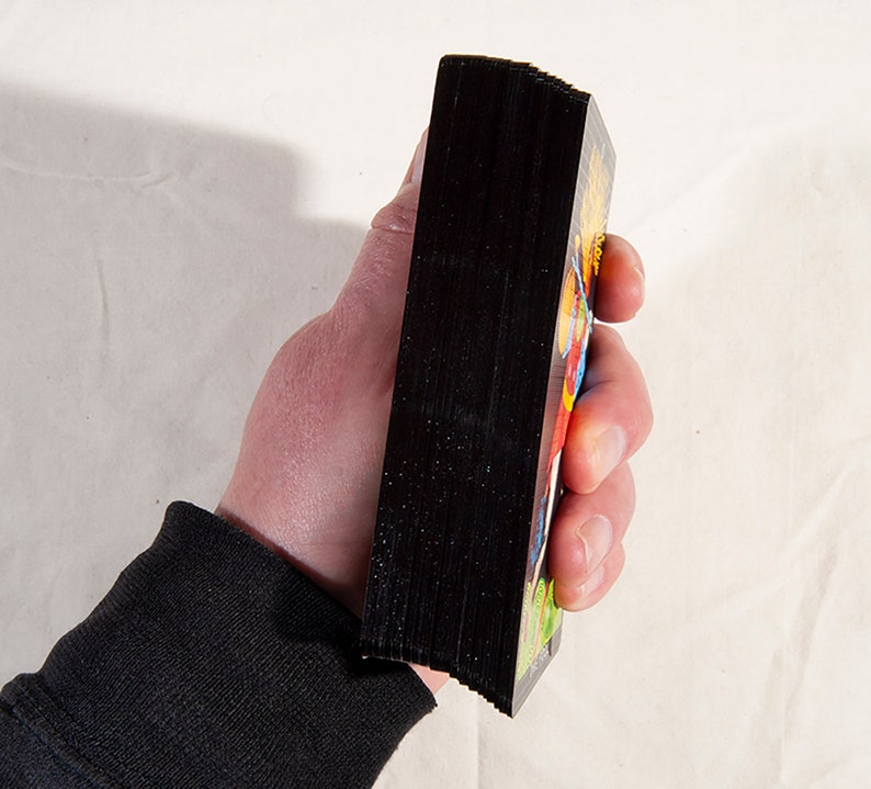 Puede incluir: Una baraja de cartas con el dorso negro y dise&ntilde;os coloridos en el frente. Las cartas est&aacute;n apiladas y se sostienen en una mano.