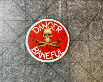 Puede incluir: Una pegatina redonda roja y blanca con las palabras "Danger Baneful" en letras blancas. Un cráneo y huesos cruzados están en el centro de la pegatina.