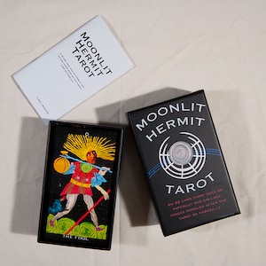 Puede incluir: Una caja negra con el texto "Moonlit Hermit Tarot" y un dise&ntilde;o circular. La caja contiene una baraja de 82 cartas de tarot con im&aacute;genes de recortes de papel y collage inspiradas en el Tarot de Marseille.