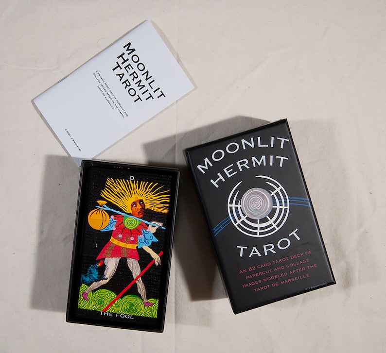 Puede incluir: Una caja negra con el texto "Moonlit Hermit Tarot" y un dise&ntilde;o circular. La caja contiene una baraja de 82 cartas de tarot con im&aacute;genes de recortes de papel y collage inspiradas en el Tarot de Marseille.