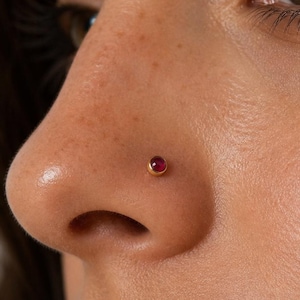 May include: A small, round, gold-toned nose stud featuring a smooth, deep red gemstone set in the center. The jewelry is positioned on the side of the nostril, showcasing a minimalist design with a polished metal finish and a vibrant red stone.