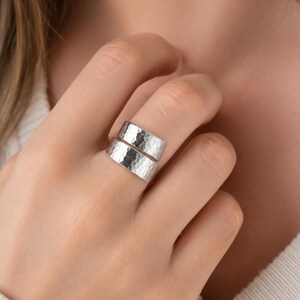 May include: A silver-toned bypass ring featuring a wide, open-ended band design. The surface of the metal is finished with a distinct hammered texture, creating a series of small, reflective indentations across the entire outer face of the ring.