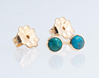May include: A pair of gold stud earrings featuring round, polished turquoise cabochons set in gold bezels. The earrings include matching gold flower-shaped earring backs, which are positioned separately from the main stud components.
