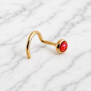 May include: A gold-colored nose ring with a curved post and a round, red opal stone. The jewelry is set against a white and gray marble background. The opal has a speckled appearance.