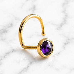 May include: A gold-toned nose ring featuring a curved post with a small ball end and a circular setting holding a faceted, deep purple gemstone. The jewelry piece has a polished metallic finish and a classic, simple design for facial piercing.