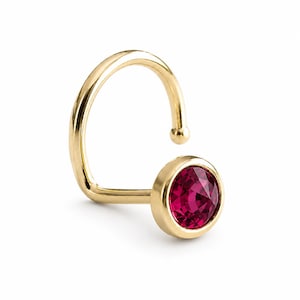 May include: A gold nose ring featuring a curved post with a small ball end and a circular setting holding a faceted, deep pink gemstone. The metal has a polished finish, and the stone is securely mounted in a smooth, rounded bezel setting.
