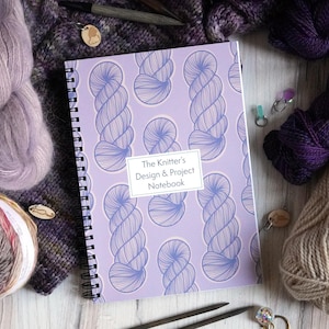 May include: A purple notebook with a spiral binding and a design of blue yarn balls on the cover. The text on the cover reads "The Knitter's Design & Project Notebook". The notebook is surrounded by balls of yarn in various colors, including purple, pink, and brown.