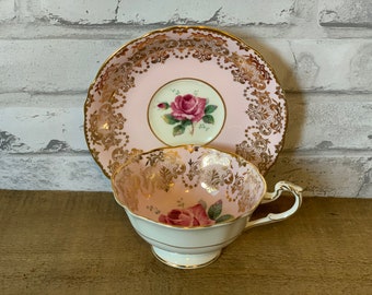 May include: A vintage teacup and saucer set. The set features a pink background with gold filigree and a rose motif. The teacup has a white base and a delicate handle. The saucer has a matching design. A classic tea set.