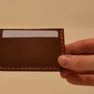 May include: A brown leather card holder with orange stitching. The card holder has a slot for a card on the top.