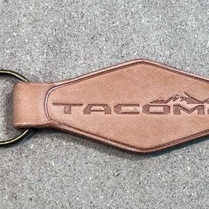 Könnte beinhalten: Hellbrauner Leder-Schlüsselanhänger mit Messingring. Der rautenförmige Anhänger trägt den Schriftzug "TACOMA" und ein Bergmotiv. Das Leder hat eine natürliche, strukturierte Oberfläche. Länge ca. 8 cm.