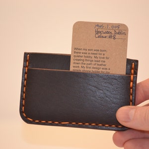 May include: A brown leather wallet with orange stitching and a brown card inside. The card has handwritten text that reads "MWS.1.005 Horween Dublin Color #8" and a short paragraph about the maker's inspiration for creating the wallet.