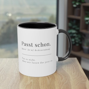 Peut inclure: Mug en céramique blanche avec intérieur et anse noirs. Le mug présente l'expression allemande "Passt schon" en texte noir, ainsi que du texte supplémentaire en dessous. Le mug est posé sur une surface en bois.