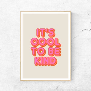 May include: A framed art print with the words "IT'S COOL TO BE KIND" in bubble letters. The text is in bright pink with an orange outline, set against a beige background. The frame is a light wood color.