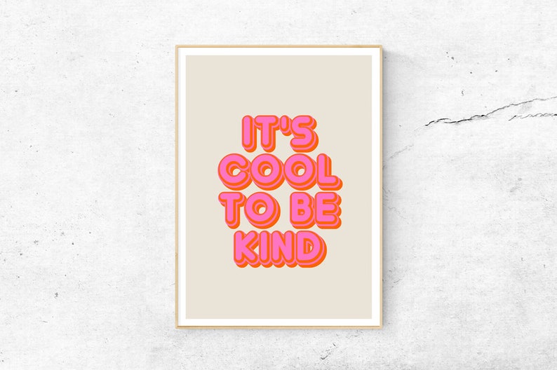 May include: A framed art print with the words "IT'S COOL TO BE KIND" in bubble letters. The text is in bright pink with an orange outline, set against a beige background. The frame is a light wood color.