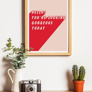 May include: A framed print with the text "HELLO! YOU'RE LOOKING GORGEOUS TODAY" in red, set against a red and beige background. The print is displayed on a wooden surface with a vintage camera, a white vase with greenery, and a small potted cactus.