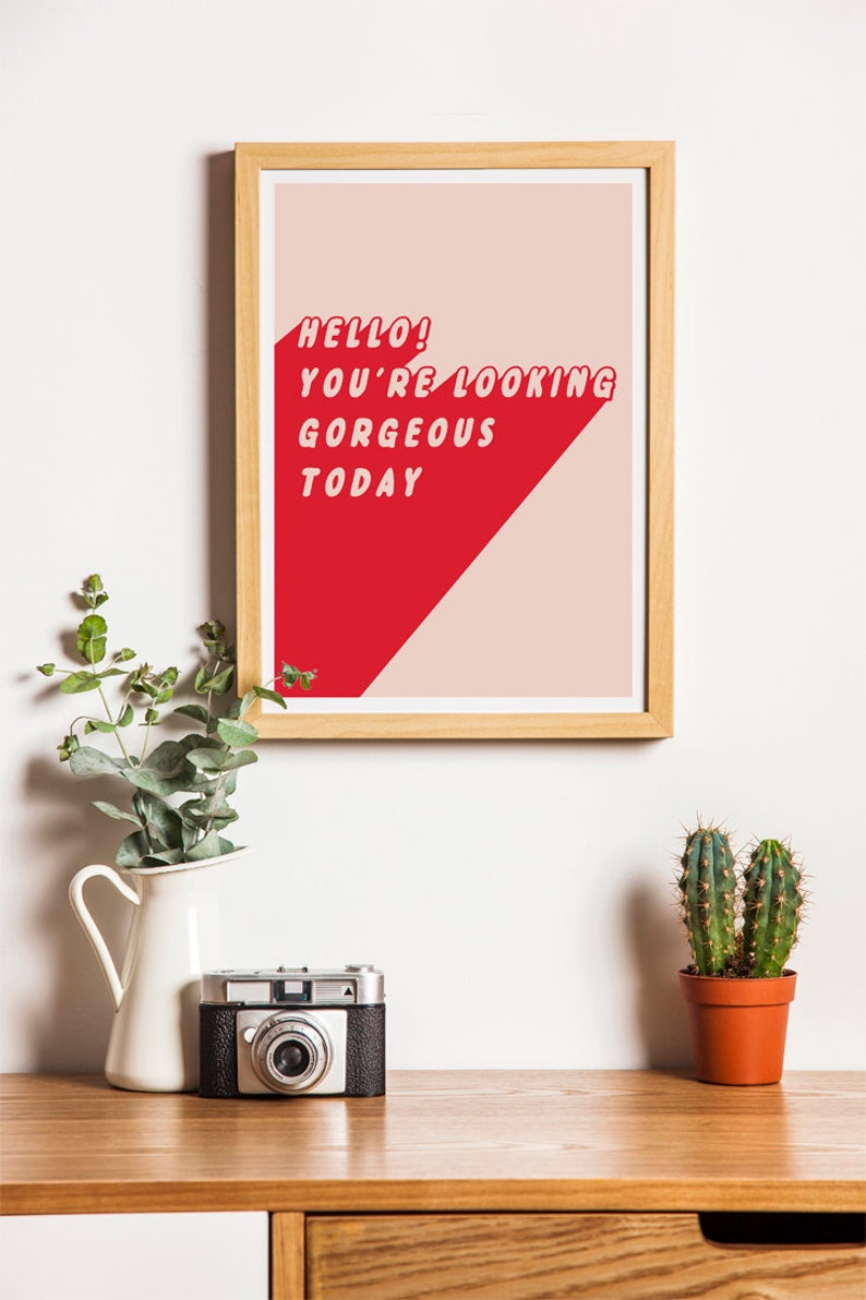 May include: A framed print with the text "HELLO! YOU'RE LOOKING GORGEOUS TODAY" in red, set against a red and beige background. The print is displayed on a wooden surface with a vintage camera, a white vase with greenery, and a small potted cactus.