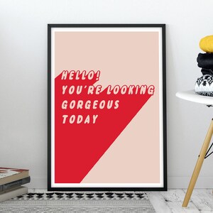 May include: A framed rectangular wall art print featuring a light pink background with a bold red diagonal shape. The text HELLO! YOU'RE LOOKING GORGEOUS TODAY is printed in white, sans-serif capital letters across the red and pink sections.