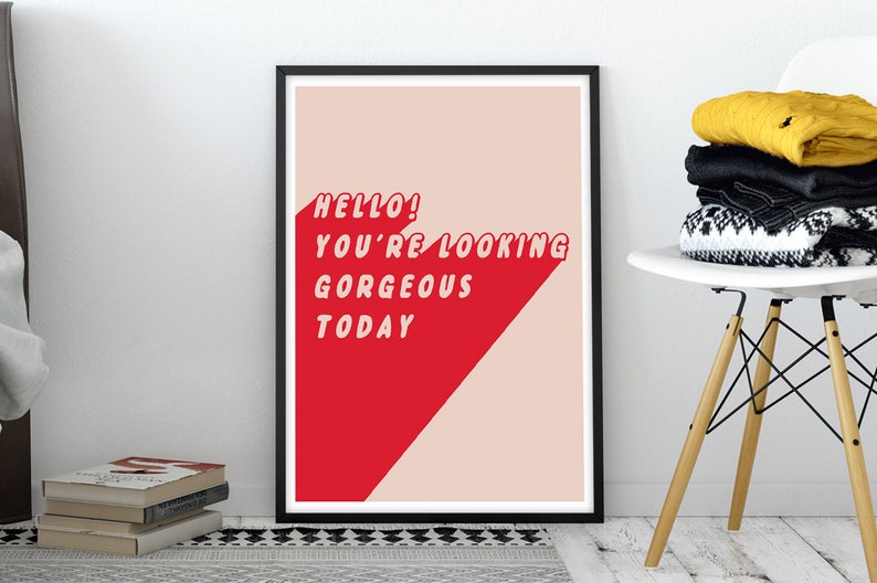 May include: A framed rectangular wall art print featuring a light pink background with a bold red diagonal shape. The text HELLO! YOU'RE LOOKING GORGEOUS TODAY is printed in white, sans-serif capital letters across the red and pink sections.