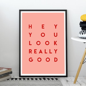 Pu&ograve; includere: Una stampa artistica incorniciata con sfondo corallo e la scritta rossa "HEY YOU LOOK REALLY GOOD". La stampa &egrave; in una cornice nera ed &egrave; appoggiata a una parete bianca. Sono visibili anche una pila di libri e una sedia bianca con vestiti piegati.