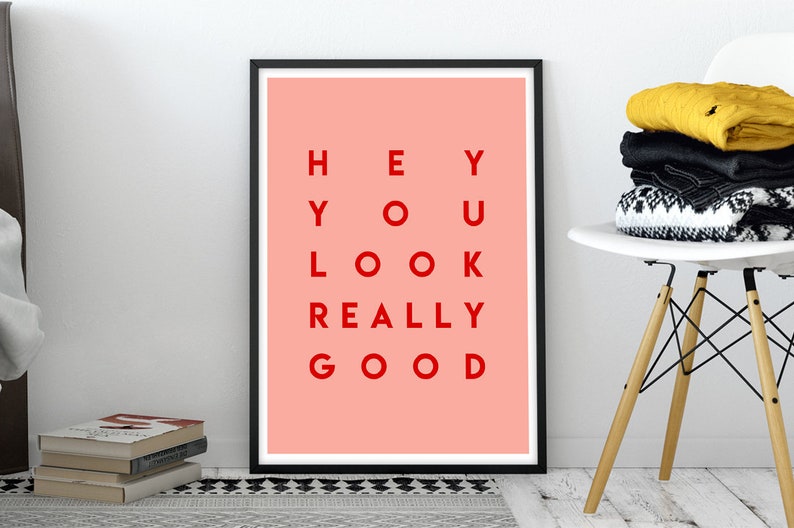 Pu&ograve; includere: Una stampa artistica incorniciata con sfondo corallo e la scritta rossa "HEY YOU LOOK REALLY GOOD". La stampa &egrave; in una cornice nera ed &egrave; appoggiata a una parete bianca. Sono visibili anche una pila di libri e una sedia bianca con vestiti piegati.
