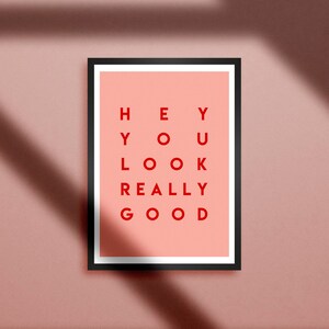 Pu&ograve; includere: Una stampa artistica incorniciata con la scritta "HEY YOU LOOK REALLY GOOD" in lettere rosse su sfondo pesca. La cornice nera &egrave; rettangolare. La stampa &egrave; esposta su una parete rosa con ombre.