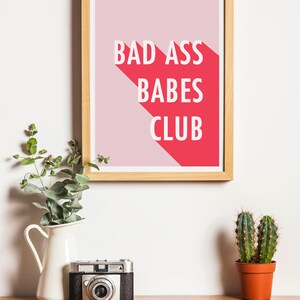 May include: A framed print with the text "BAD ASS BABES CLUB" in white letters on a pink and red background. The print is displayed on a wooden surface with a vintage camera, a white vase with greenery, and a small potted cactus.