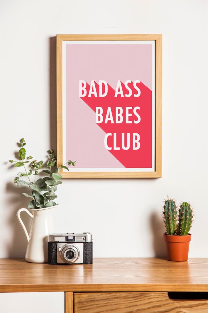 May include: A framed print with the text "BAD ASS BABES CLUB" in white letters on a pink and red background. The print is displayed on a wooden surface with a vintage camera, a white vase with greenery, and a small potted cactus.