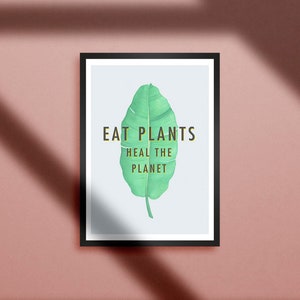 K&ouml;nnte beinhalten: Gerahmter Kunstdruck mit schwarzem Rahmen. Der Druck zeigt ein gro&szlig;es gr&uuml;nes Blatt mit den Worten "EAT PLANTS HEAL THE PLANET" in goldfarbenen Buchstaben. Der Hintergrund ist hellblau.