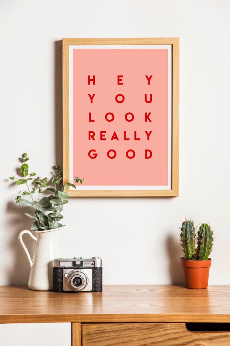 Pu&ograve; includere: Una stampa incorniciata con la scritta "HEY YOU LOOK REALLY GOOD" in lettere rosse su sfondo rosa. Una fotocamera vintage, un piccolo cactus in un vaso di terracotta e un vaso bianco con piante verdi sono su una superficie di legno.