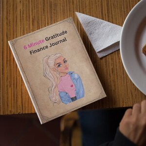 May include: A brown journal with the text "6 Minute Gratitude Finance Journal" on the cover. The cover also features a cartoon illustration of a woman with blonde hair wearing a pink shirt and a blue denim jacket.