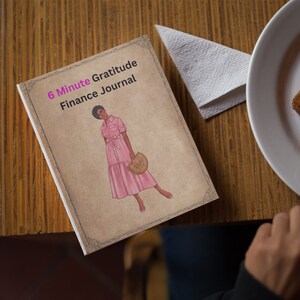 May include: A journal titled "6 Minute Gratitude Finance Journal" with a woman in a pink dress illustration. The journal is on a wooden table next to a napkin and a plate. The journal's cover has a vintage design.