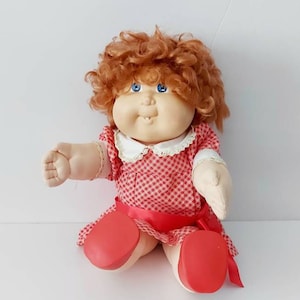 May include: A red-haired Cabbage Patch Kid doll wearing a red and white checkered dress with a red ribbon. The doll has blue eyes and a big smile.