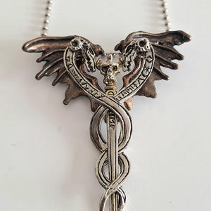 Peut inclure: Collier pendentif argenté avec un motif détaillé de dragons ailés, d'un crâne et d'une épée. Le pendentif est suspendu à une chaîne fine. Le collier a un thème gothique ou fantastique.