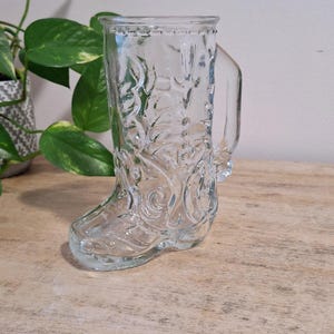 Puede incluir: Una taza transparente en forma de bota con asa. La taza tiene diseños en relieve de flores y volutas. Se encuentra sobre una superficie de madera, con una planta en el fondo.