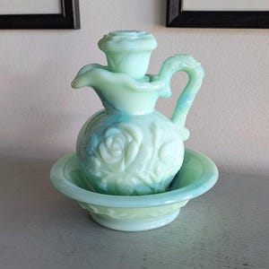 Op de afbeelding: Een vintage groene glazen zeepdispenserset met een kan en een kom. De kan heeft een bloemenmotief en een handvat. De kom is rond en heeft een gegolfde rand.