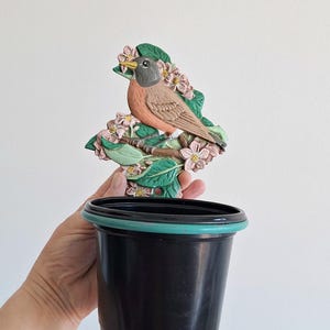 以下が含まれることがあります： 緑色の縁が付いた黒いプラスチック製の植木鉢。装飾的な鳥と花のアクセントが鉢の上部に取り付けられています。鳥は茶色、灰色、オレンジ色で、ピンクの花が咲く枝にとまっています。