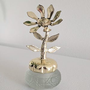 Op de afbeelding: Een decoratieve parfumfles met een bloemvormige bovenkant. De bloem is gemaakt van goudkleurig metaal en heeft gedetailleerde bloemblaadjes en bladeren. De fles is transparant met een matte afwerking.