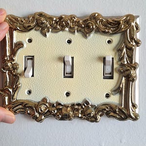 May include: A decorative light switch plate with a gold ornate frame and three white toggle switches.
