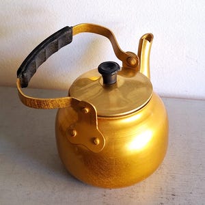 Puede incluir: Una tetera dorada con un asa y un pomo de tapa negros. La tetera tiene un cuerpo redondeado, un pico y un asa que se arquea hacia arriba. La tetera es de metal y parece vintage.