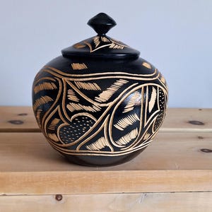 Peut inclure: Un pot rond en bois noir avec un couvercle, orné de motifs complexes de feuilles et de fleurs sculptées dans un bois plus clair. Le pot est présenté sur une surface en bois.