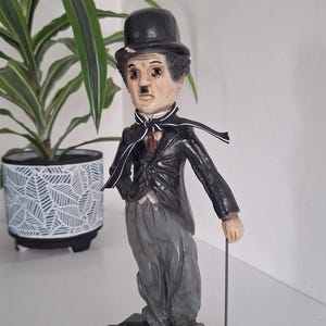 Puede incluir: Figura de Charlie Chaplin con su atuendo icónico: bombín negro, traje y bastón. La figura tiene un bigote pintado y está sobre una base gris. Una planta en una maceta estampada está en el fondo.