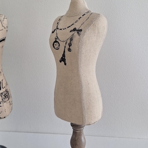 Op de afbeelding: Een beige stoffen paspop met een houten voet en bovenkant. De paspop heeft een zwarte illustratie van een ketting met een klok, Eiffeltoren en andere bedels. Het is een decoratief item.