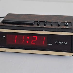 May include: A vintage Cosmo alarm clock displaying 11:21 PM in red digital numbers. The clock has a black face with gold trim and a wood-grain top. It features alarm and time setting buttons.