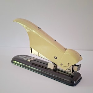 Puede incluir: Una grapadora vintage amarilla con base negra. La grapadora tiene un diseño curvo único y un mecanismo de grapado de metal.