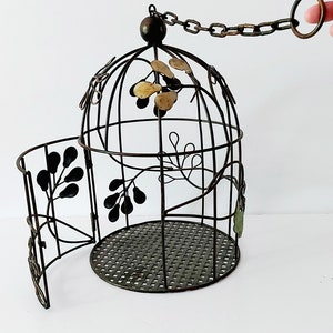 May include: A black metal birdcage with a decorative floral design. The cage has a chain attached to the top and a door that opens on the side.