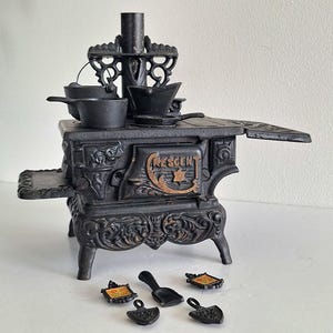 May include: A miniature black cast iron toy stove with ornate scrollwork detailing, featuring the word CRESCENT embossed on the front door. The stove includes a chimney, side shelves, and several small black metal cooking accessories and tools.