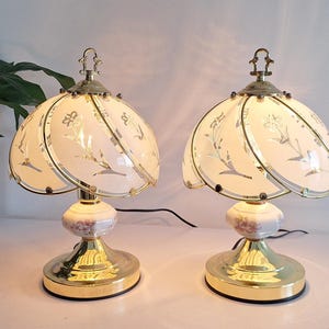 Pode incluir: Duas luminárias de mesa combinando com abajures creme com design floral. As luminárias têm base e detalhes dourados, com um remate decorativo. A base tem um aspeto marmoreado. Uma planta em vaso está no fundo.