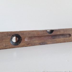 Könnte beinhalten: Eine rechteckige Wasserwaage aus Holz mit verwitterter brauner Oberfläche, einer kreisförmigen Glaslibellenfassung auf der linken Seite und einer horizontalen vertieften Rinne an der Oberkante, die eine abgenutzte Textur und sichtbare Holzmaserung aufweist.